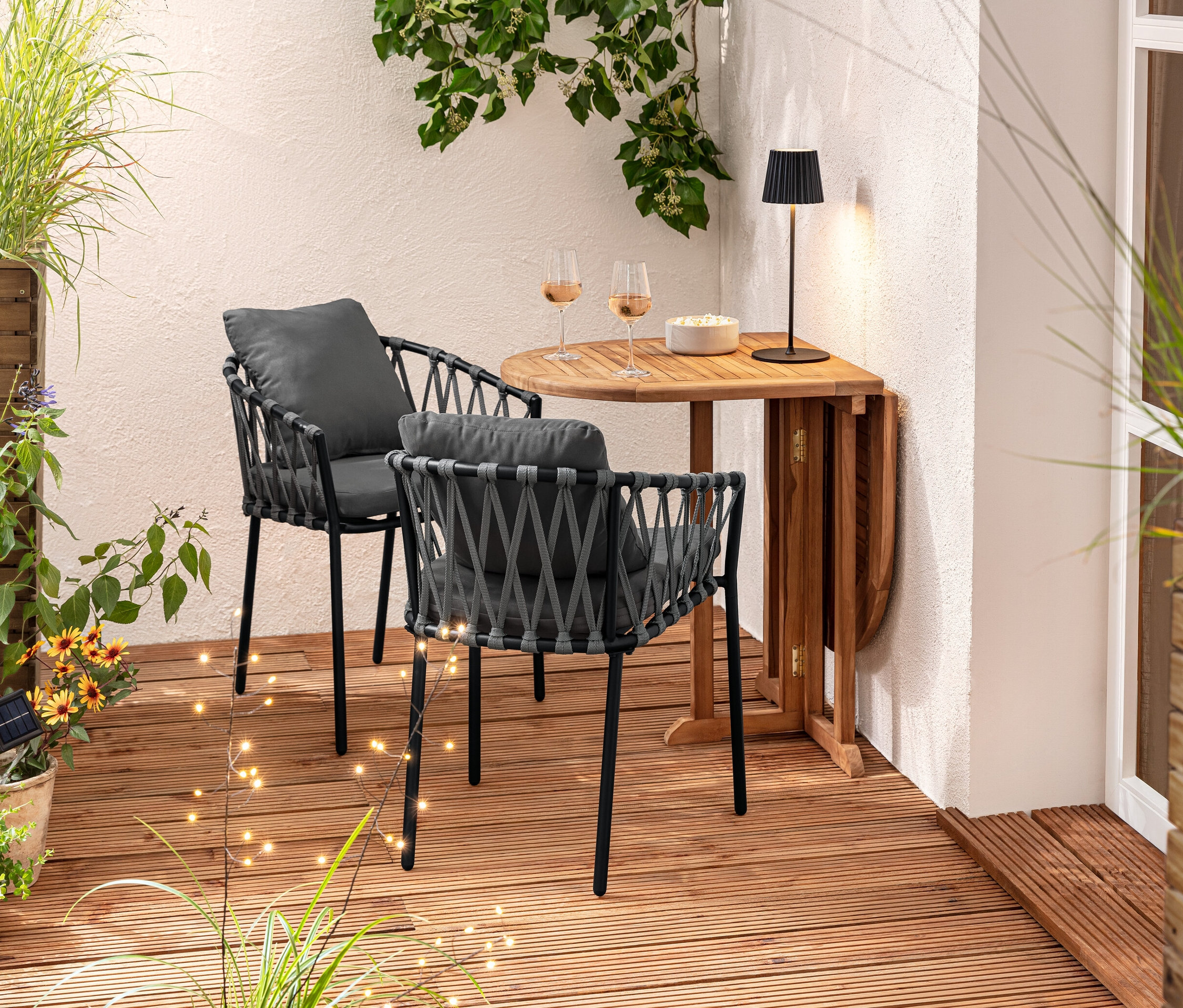 Zwei Dining-Sessel mit Textilgeflecht stehen an einem Holztisch. Auf dem Tisch stehen Gläser und eine Schüssel. Im Hintergrund befindet sich ein Pflanzkasten-Raumteiler und 3 Solar-Spieße.