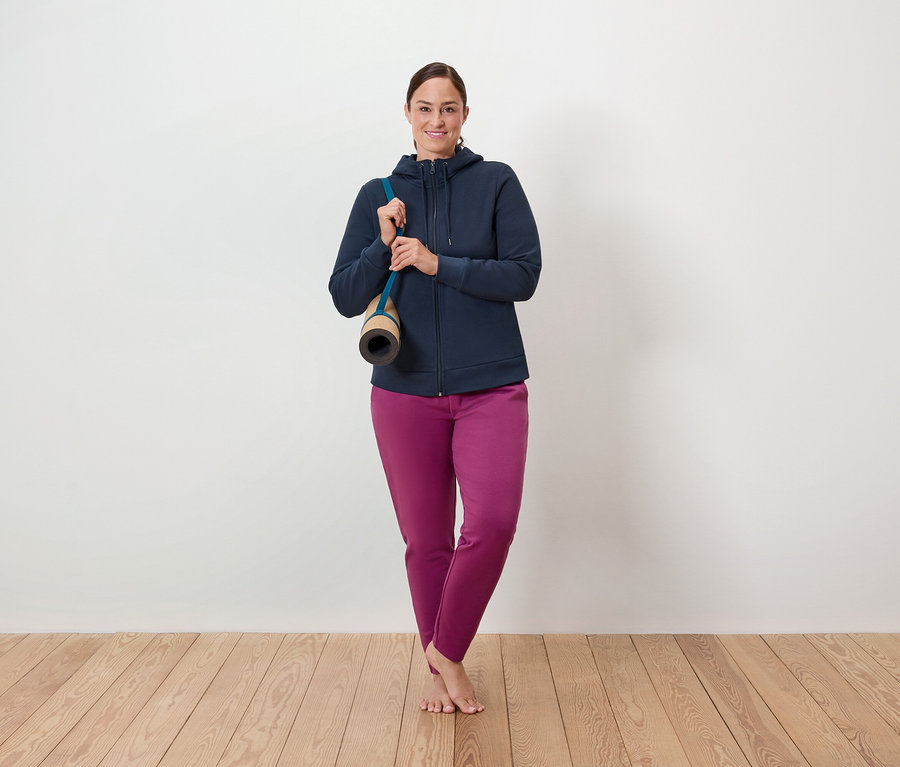Eine Frau steht auf einem Holzboden, eine Yogamatte über die Schulter gehängt, und trägt eine blaue Reißverschlussjacke und eine violette Hose.