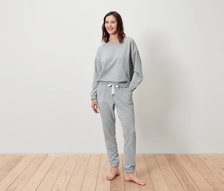 Frau steht und lächelt, sie trägt einen grauen Pullover und eine Jogginghose.