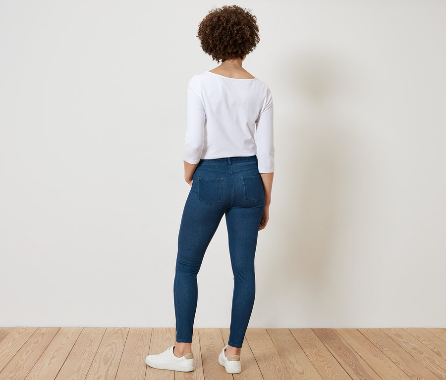 Rückansicht einer Frau mit lockigem Haar, die ein weißes Hemd und blaue Jeans trägt.