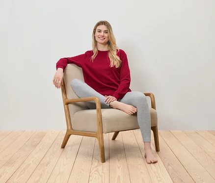 Blonde Frau sitzt in einem beigen Sessel, trägt ein rotes Sweatshirt und graue Leggings.