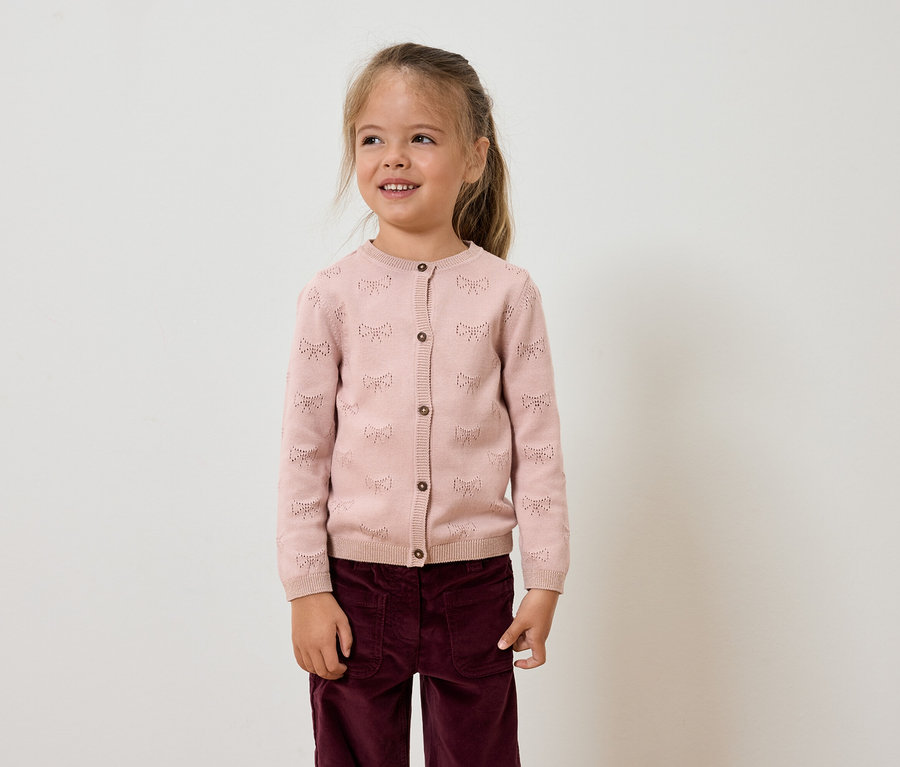 Mädchen mit braunen Haaren trägt eine rosa Strickjacke und violette Hosen.