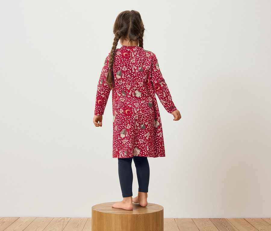 Mädchen in rotem Musterkleid und dunkelblauen Leggings steht auf einem Holzsockel.