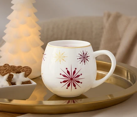 Auf einem goldenen Tablett stehen eine weiße Tasse mit roten und goldenen Schneeflocken, eine Schale mit Lebkuchen und ein leuchtender Baum.