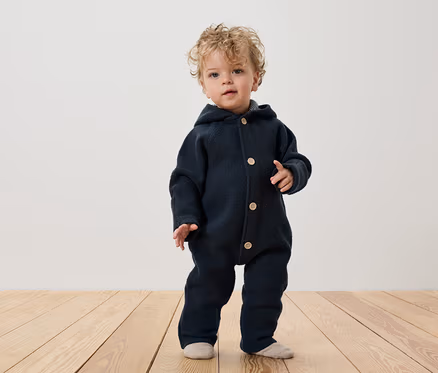 Ein Kind mit lockigem Haar steht auf einem Holzboden und trägt einen dunkelblauen Overall mit Kapuze und Knöpfen.