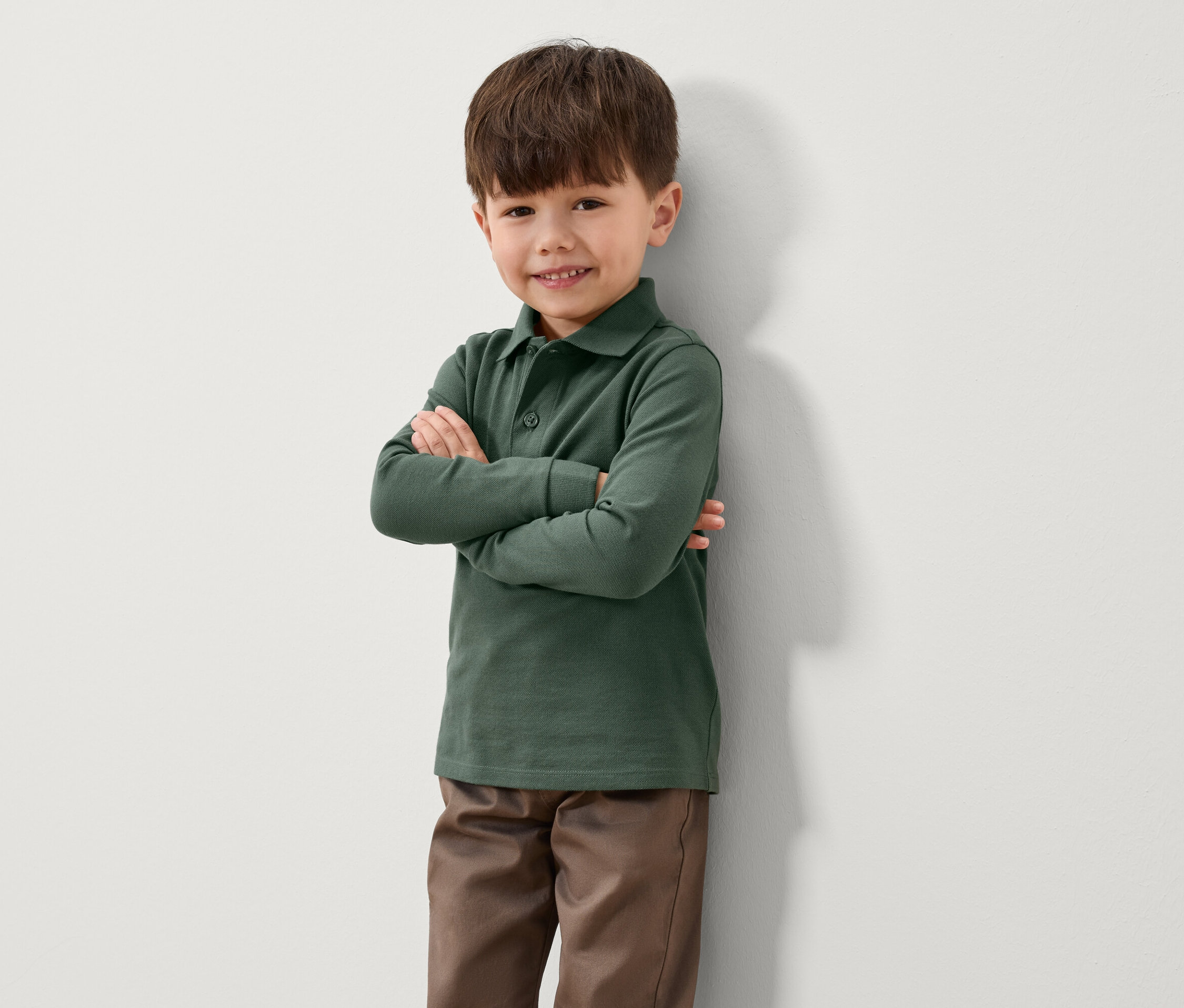 Ein Junge steht mit verschränkten Armen an einer Wand. Er trägt ein grünes Langarm-Poloshirt und braune Hosen.