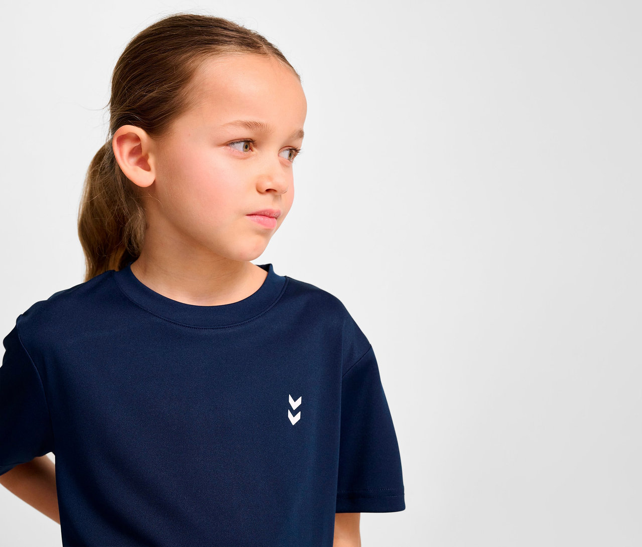Porträt eines Mädchens mit braunen Haaren, das ein dunkelblaues HUMMEL HMLJR Pulse Trainings-T-Shirt trägt.