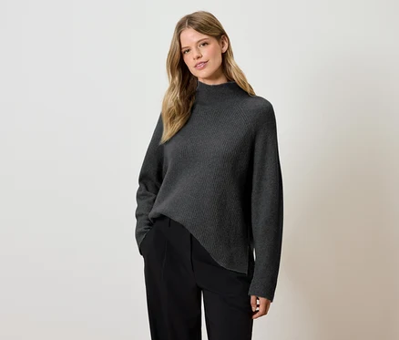 Frau posiert in einem grauen Pullover und einer schwarzen Hose.