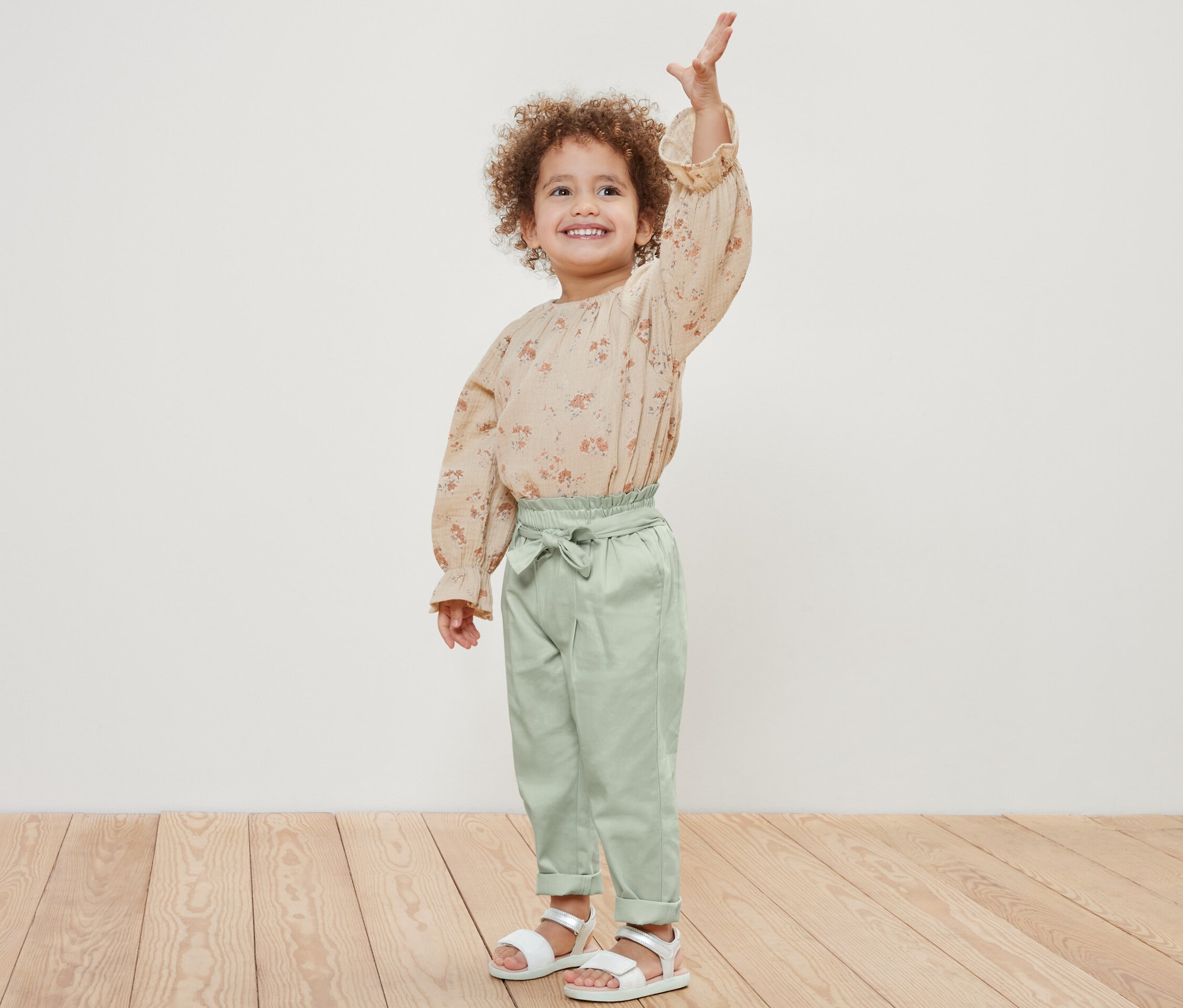 Ein lächelndes Mädchen posiert und winkt. Sie trägt eine geblümte Bluse und eine Kinder-Pull-on-Pants.