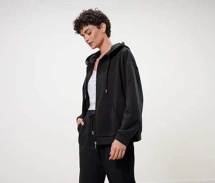 Frau mit kurzen lockigen Haaren trägt einen schwarzen Kapuzenpullover, eine schwarze Trainingshose und ein weißes Top.