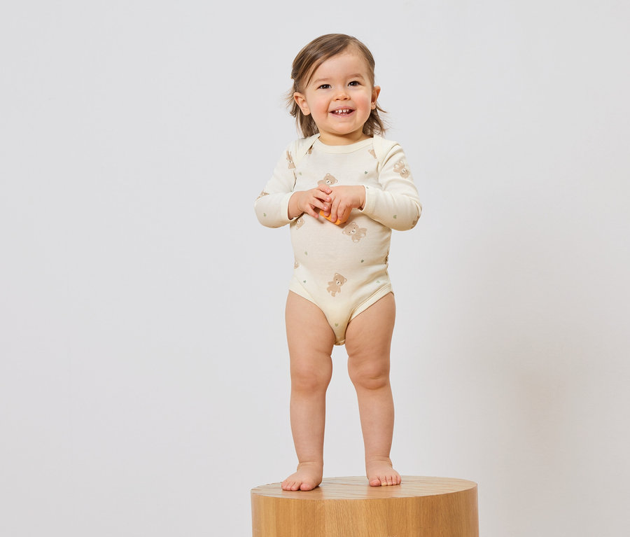 Lächelndes Kleinkind im Teddy-Body steht auf einem Hocker vor einem weißen Hintergrund.