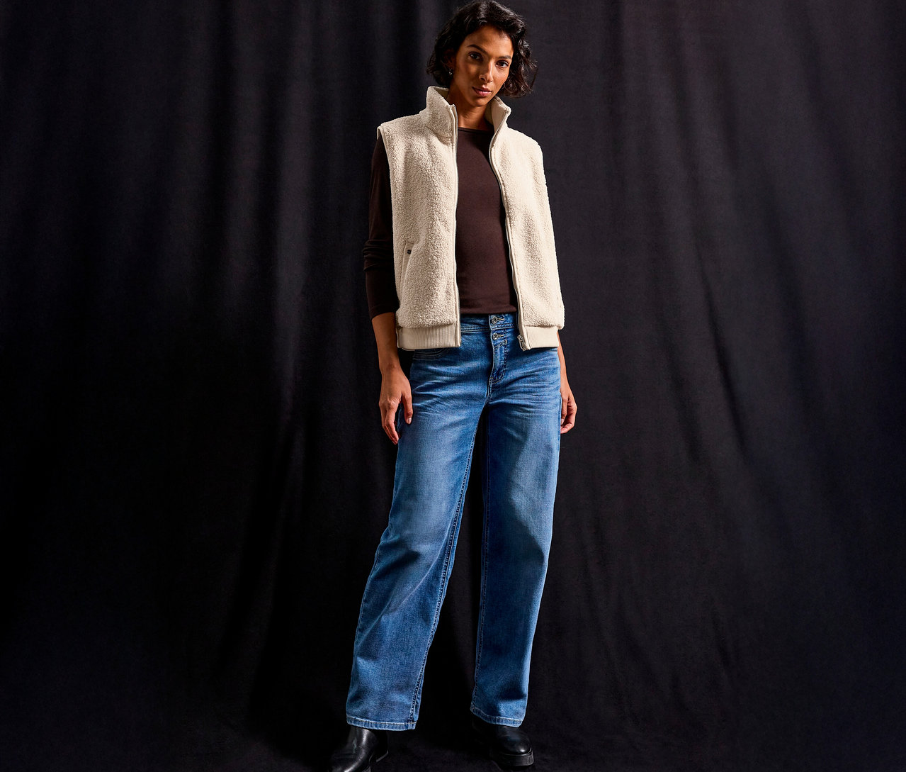 Frau trägt eine Street One Teddyweste über einem braunen Langarmshirt und Jeans vor einem schwarzen Hintergrund.