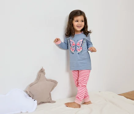 Ein kleines Mädchen mit braunen Haaren trägt einen blauen Pyjama mit einem Schmetterlingsmuster und pinkfarbenen, gestreiften Hosen. Neben ihr liegen ein sternförmiges und ein wolkenförmiges Kissen auf einer weißen Decke.