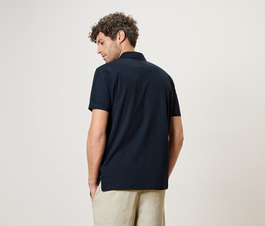 Ein Mann von hinten, trägt ein marineblaues Polohemd und eine beigefarbene Hose vor einem neutralen Hintergrund.