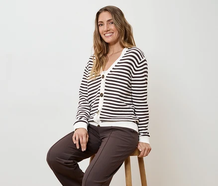 Frau mit braunen Hosen und gestreifter Strickjacke sitzt auf einem Hocker vor einem weißen Hintergrund.