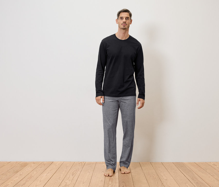 Mann steht barfuß in grauer Hose und schwarzem Langarmshirt vor einer Wand auf einem Holzboden.