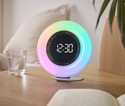 Auf einem Nachttisch steht eine bunte Lichtwecker mit der Uhrzeit 12:30, neben einem Glas Wasser und einem Buch.