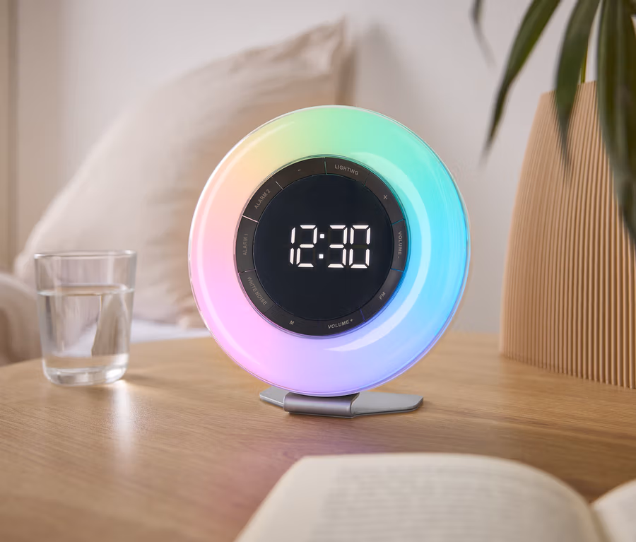 Auf einem Nachttisch steht eine bunte Lichtwecker mit der Uhrzeit 12:30, neben einem Glas Wasser und einem Buch.