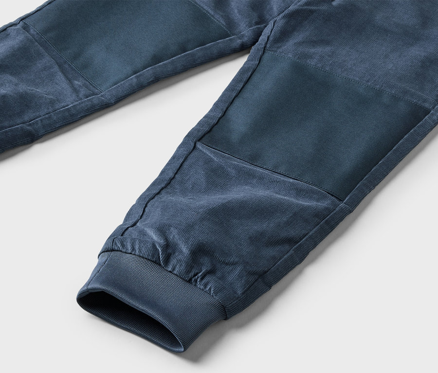 Nahaufnahme einer marineblauen Cordhose mit elastischen Bündchen und verstärkten Kniepartien auf hellgrauem Grund.