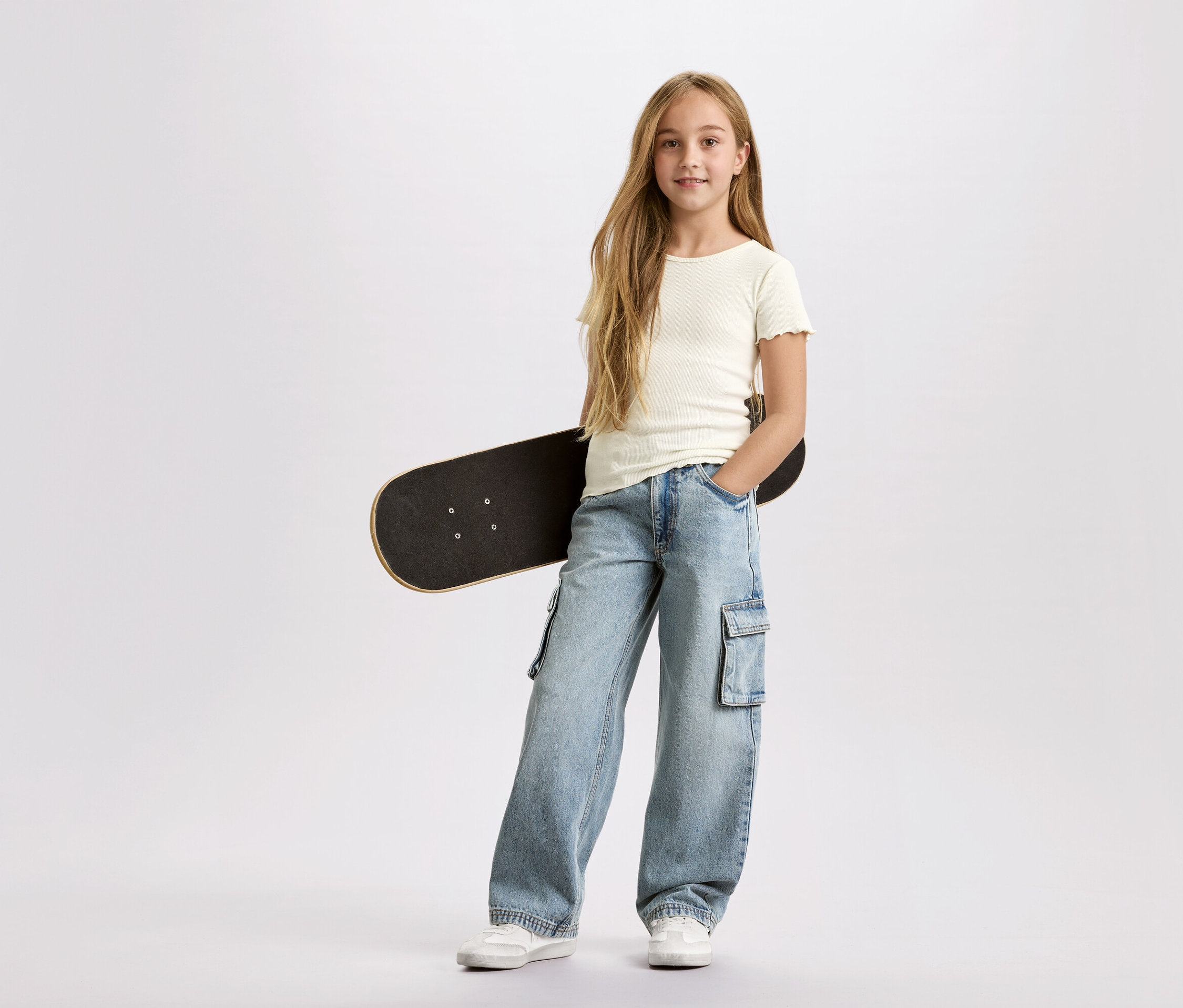 Mädchen mit langen blonden Haaren, mit einem schwarzen Skateboard unter dem Arm, trägt eine hellblaue Cargohose und ein weißes T-Shirt vor einem weißen Hintergrund.