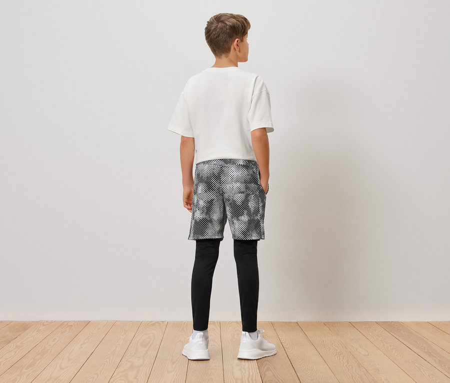 Rückansicht eines Jungen mit weißem T-Shirt, gemusterten Shorts, schwarzen Leggings und weißen Turnschuhen.
