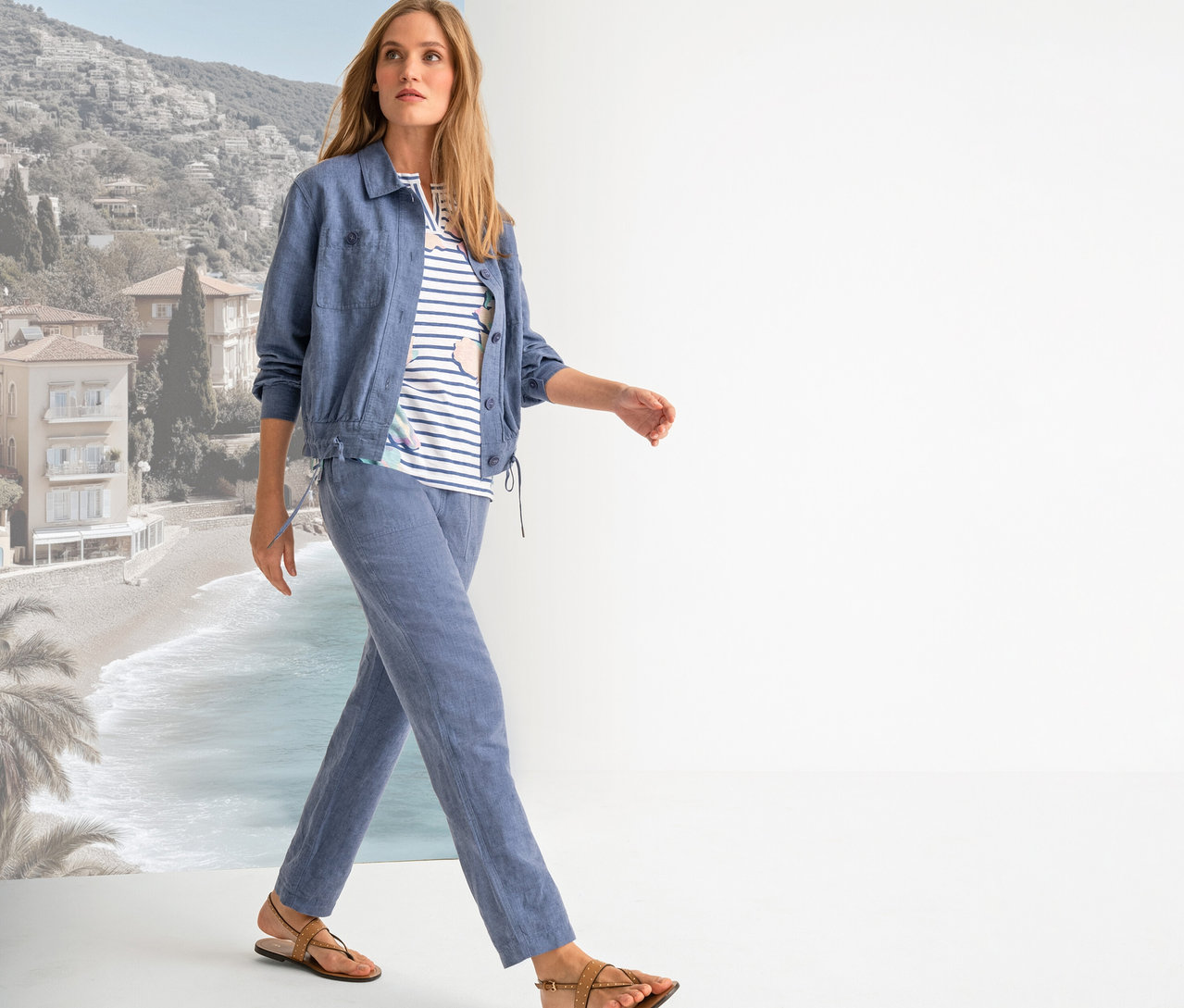 Frau geht in blauer Jacke, gestreiftem Shirt, blauer Hose und braunen Sandalen vor einem Strandhintergrund.