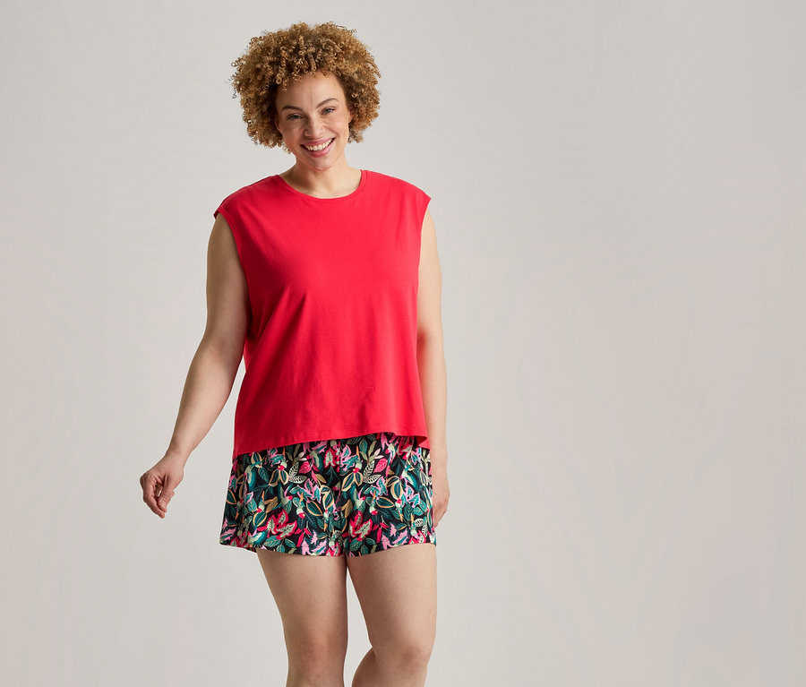 Lächelnde Frau mit lockigem Haar in einem roten ärmellosen Oberteil und kurzen, geblümten Shorts in Grün, Pink und Blau.