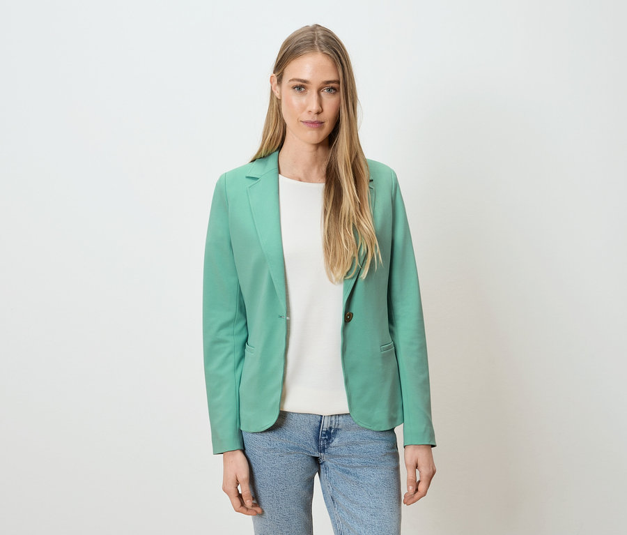 Frau trägt grünen Blazer, weißes Shirt und blaue Jeans vor einem weißen Hintergrund.