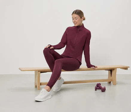 Frau sitzt auf Bank in weinrotem Sportoutfit mit Zipper-Top, Leggings und weißen Turnschuhen. Hanteln stehen auf dem Boden.