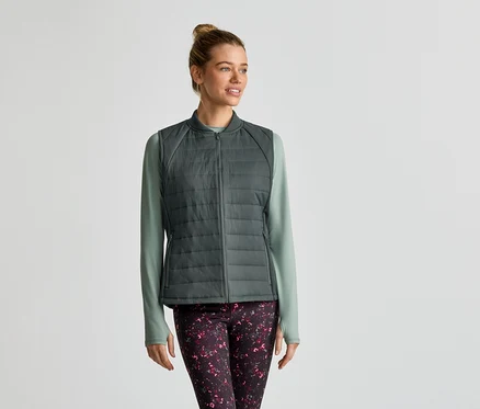 Frau mit Dutt trägt eine graue Steppweste über einem hellgrünen Langarmshirt und gemusterte Leggings.