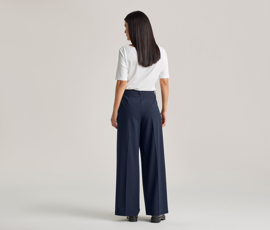 Rückansicht einer Frau mit langen, dunklen Haaren, die ein weißes T-Shirt und eine weite, marineblaue Hose mit Bund trägt.