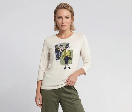 Blonde Frau trägt cremefarbenes Langarmshirt mit grafischem Druck einer Frau in gelber Jacke und olivgrüne Hose.