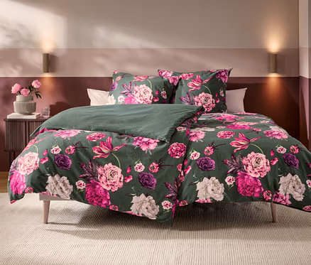 Bett mit dunkelgrüner Bettwäsche, die mit großen rosa und violetten Blumenmustern verziert ist. Eine Seite der Decke ist schlicht dunkelgrün. Daneben steht ein Nachttisch mit rosa Blumen in einer Vase.