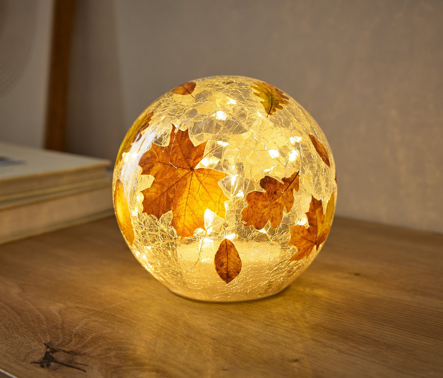 Eine kugelförmige Lampe mit rissiger Glasstruktur, verziert mit Herbstblättern, leuchtet warm auf einer Holzoberfläche. Kleine Lichter im Inneren beleuchten die Blätter.