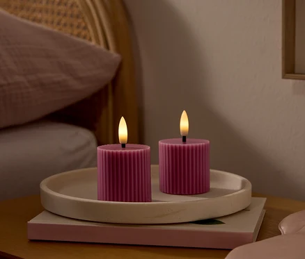 Zwei pinkfarbene, gerippte LED-Kerzen mit leuchtenden Dochten stehen auf einem hellen Tablett auf einem Nachttisch. Daneben liegt eine rosa Schlafmaske. Im Hintergrund ist ein Bett mit einem Flechtkopfteil und einem rosa Kissen zu sehen.