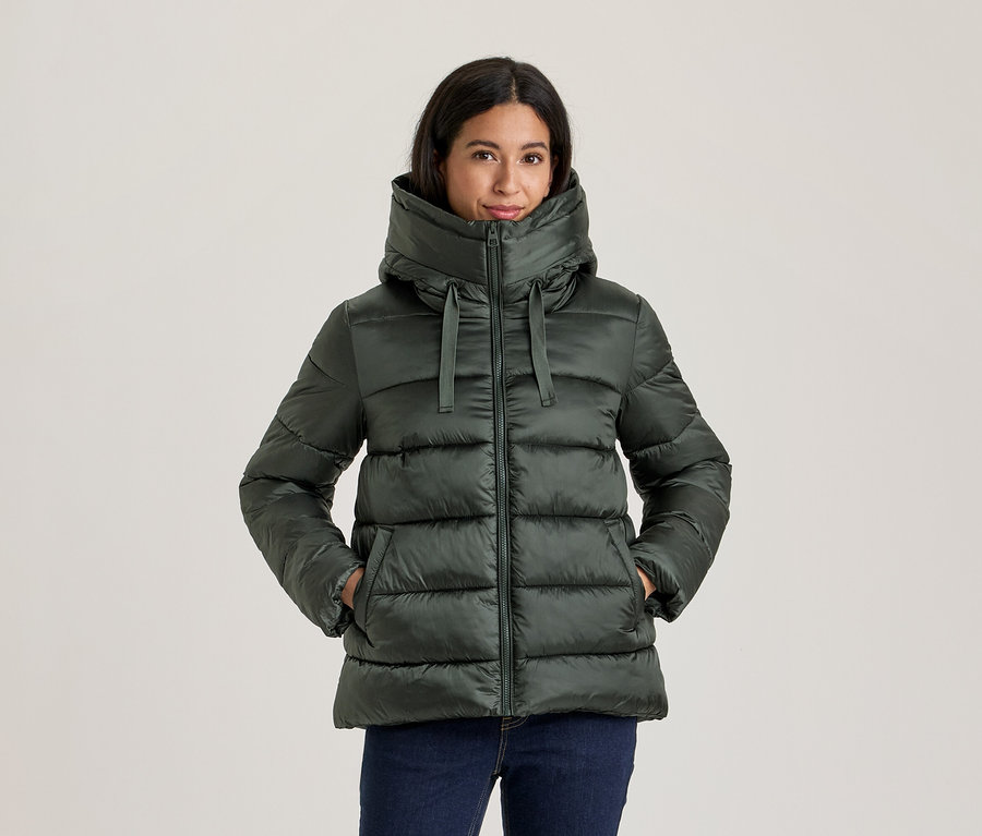 Frau in dunkelgrüner Steppjacke mit hohem Kragen und Kapuze, Hände in den Taschen, dazu dunkle Jeans.