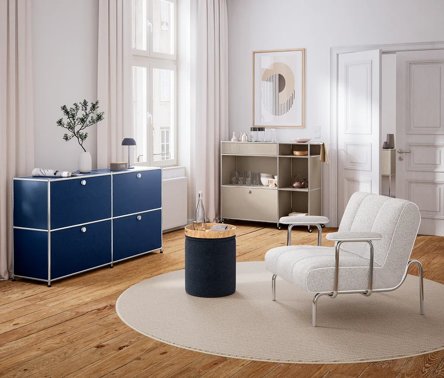 Modernes Wohnzimmer mit Holzboden und weißen Wänden. Links steht ein dunkelblaues Sideboard unter einem Fenster, darauf eine Vase und eine Lampe. In der Mitte liegt ein runder Teppich mit einem zylindrischen Beistelltisch und einer Wasserflasche. Rechts steht ein heller Sessel und ein beiges Rollregal mit Geschirr. An der Wand hängt ein abstraktes Bild.