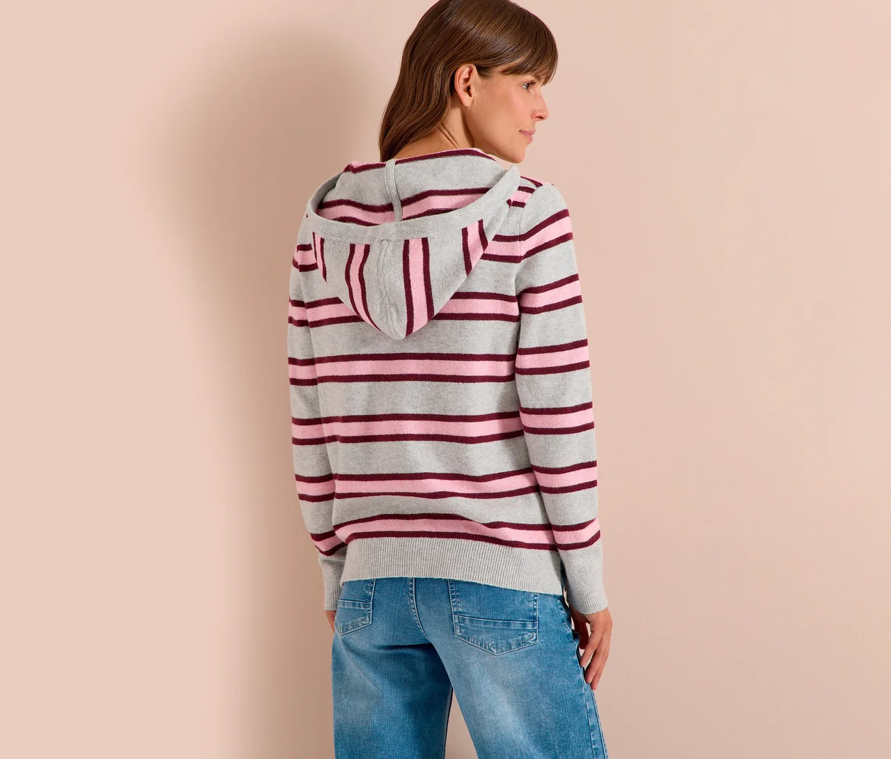 Frau von hinten, leicht zur Seite gedreht, trägt einen gestreiften CECIL Strick-Hoodie in Grau, Pink und Burgunderrot sowie blaue Jeans.
