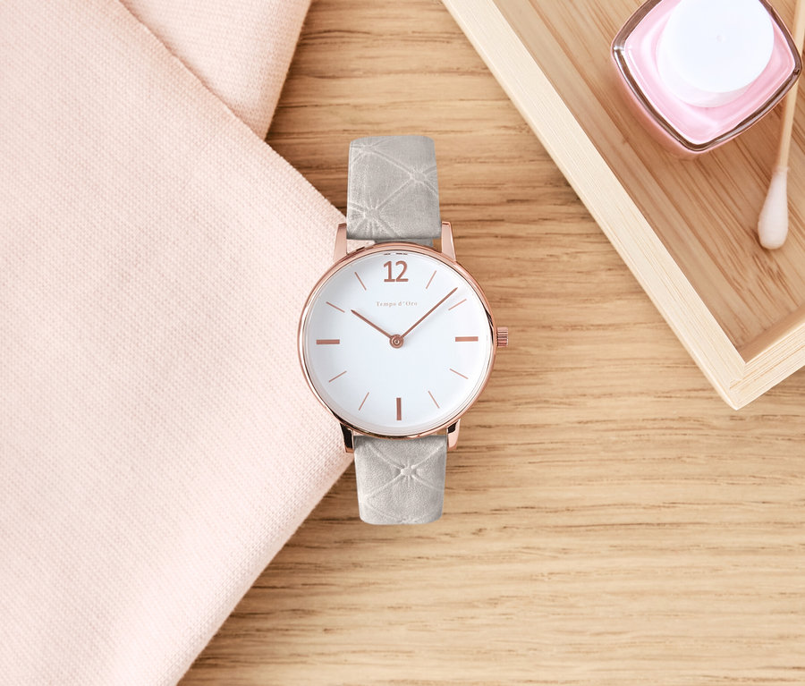 Eine Armbanduhr mit Lederband liegt auf einem Holztisch neben einem rosa Tuch und Kosmetika.