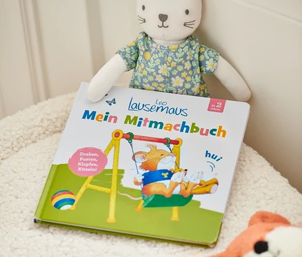 Buch »Leo Lausemaus – mein Mitmachbuch« mit einem Katzenkuscheltier und anderem Spielzeug auf einer zotteligen Oberfläche.