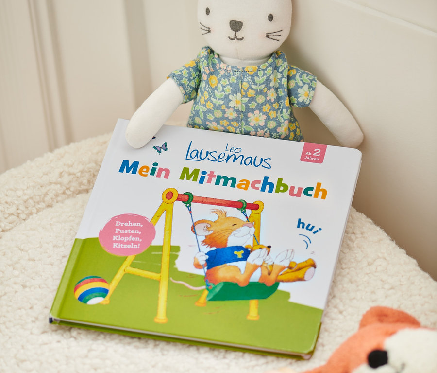 Buch »Leo Lausemaus – mein Mitmachbuch« mit einem Katzenkuscheltier und anderem Spielzeug auf einer zotteligen Oberfläche.
