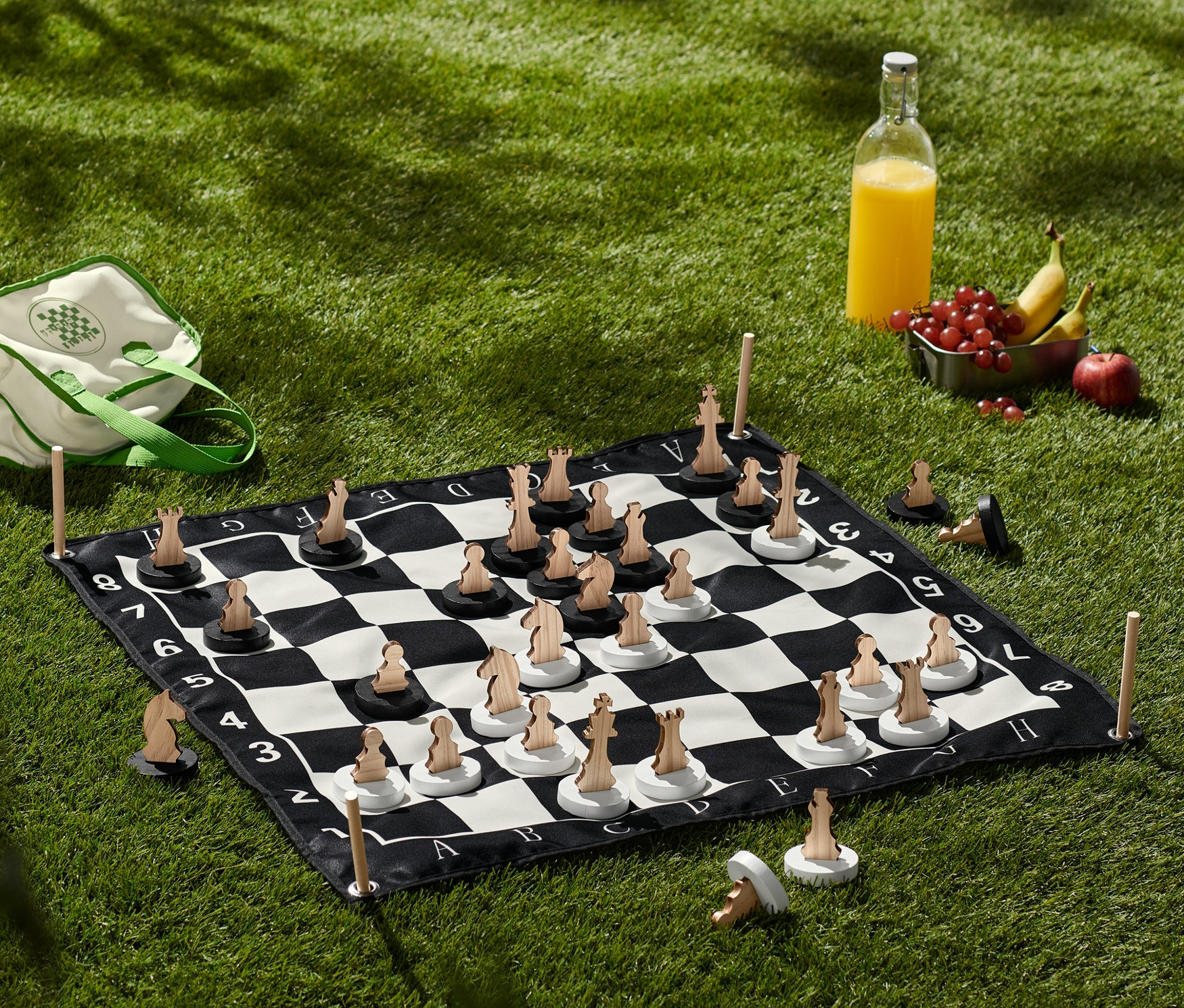 Auf dem Rasen liegt ein XL-Schachspiel mit Holzfiguren. Daneben liegen eine Tasche, Saft und Obst.