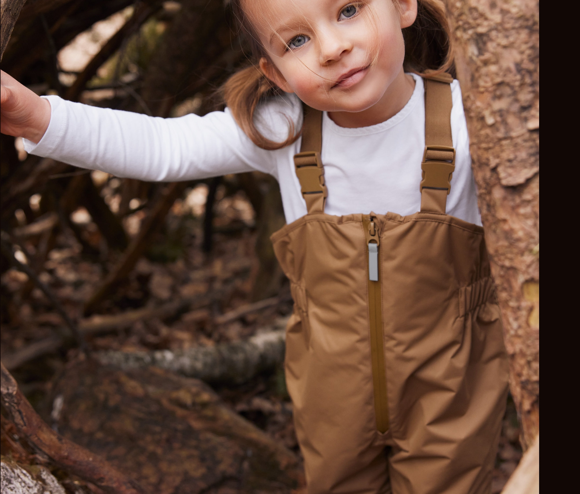 Mädchen in brauner Regenhose steht an einem Baum.