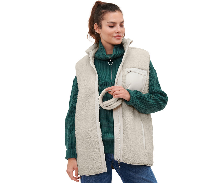 Frau hält Ohrenschützer und trägt einen grünen Pullover und eine creme Weste.