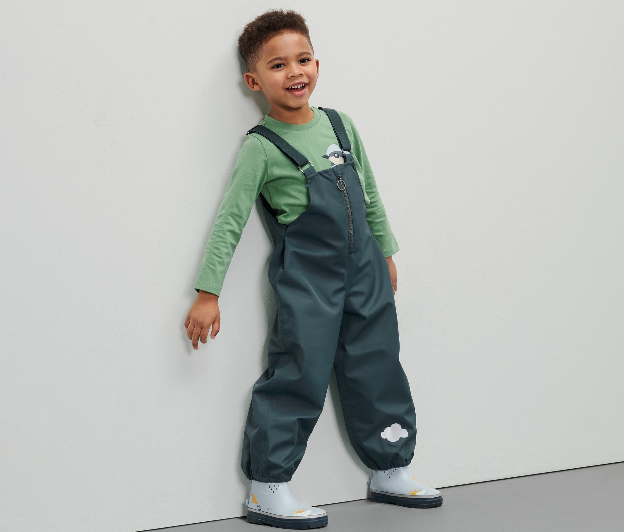 Ein Junge in einer Kleinkind-Regenhose lehnt an einer Wand.