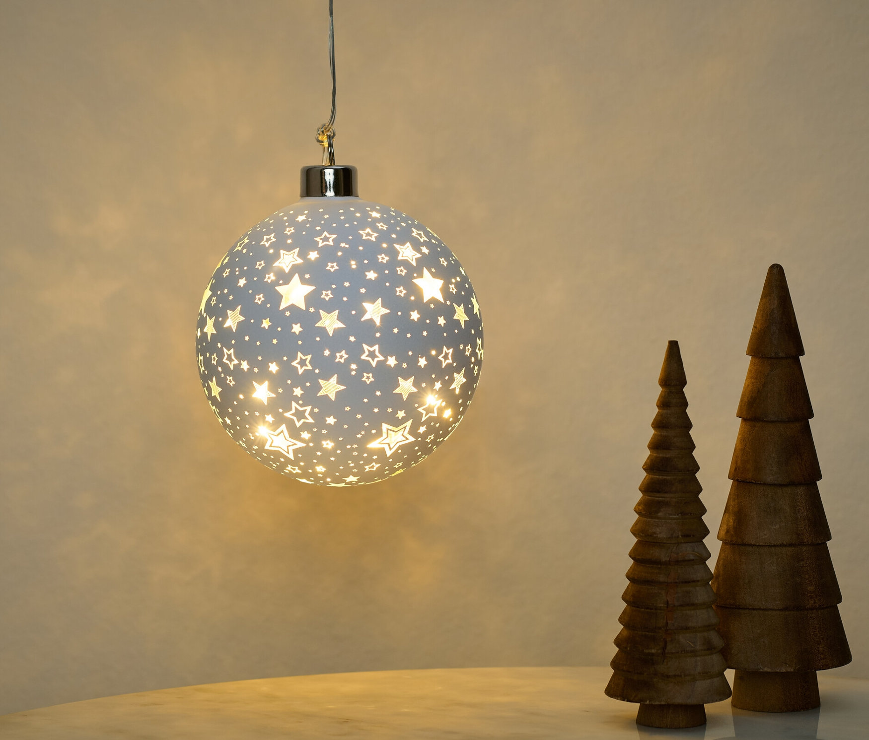 Eine hängende LED-Glaskugel mit Sternmuster und drei hölzerne Bäume auf einem Tisch.