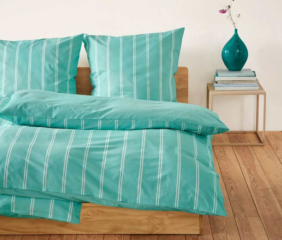 Renforcé-Bettwäsche mit blau-weißen Streifen auf einem Bett. Neben dem Bett steht ein Tisch mit einer Vase.
