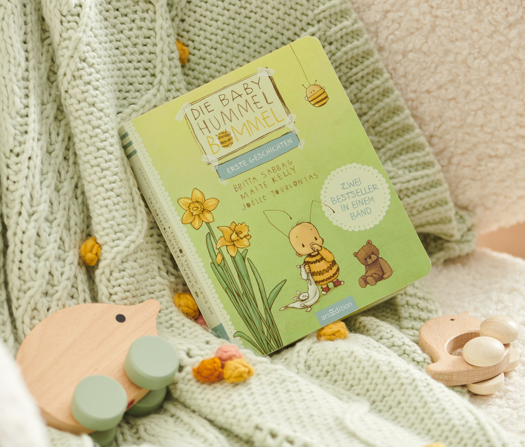 Das Buch »Baby Hummel Bommel – erste Geschichten« liegt auf einer Strickdecke mit Bommeln und Holzspielzeug.
