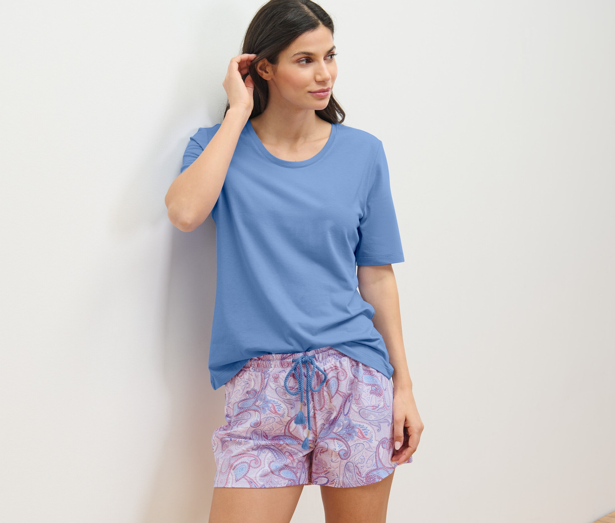 Frau in Shorty-Pyjama-Set lehnt an der Wand.