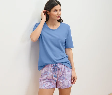Frau in Shorty-Pyjama-Set lehnt an der Wand.